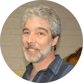 Smiling man with graying hair and beard, wearing a blue striped shirt. He stands in front of a stone wall and ornate wooden furniture.