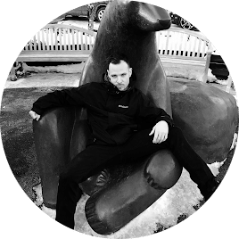 A man in a black jacket sits casually on a large, sculpted hand outdoors. A bench and snowy patches are visible in the background.