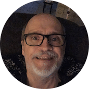 A smiling man with glasses and a gray goatee looks directly at the viewer. He is wearing a dark shirt and is sitting on a dark surface. The background is slightly blurred, focusing attention on his friendly expression.
