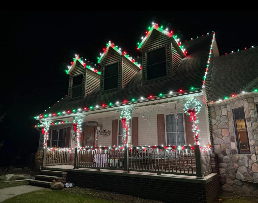 Festive home exterior at night, decorated with red, green, and white Christmas lights outlining the roof and dormers. Wreaths with red bows adorn the porch columns, creating a warm, holiday ambiance.