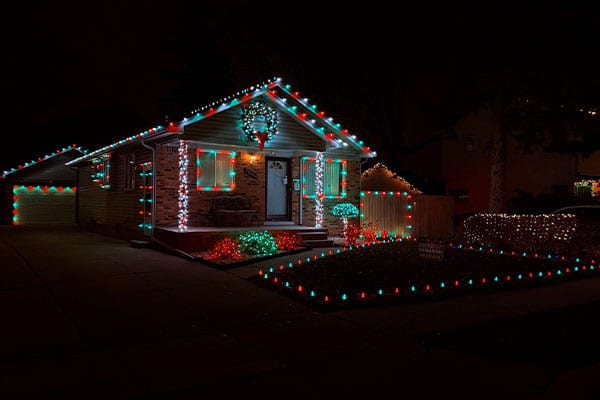 House decked out in Christmas lights at night. Red, green and white lights outline the roof, windows, and landscaping. A wreath hangs on the front door. Festive holiday scene.