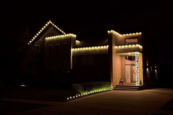 House exterior at night, decorated with warm white Christmas lights outlining the roofline, landscaping, and porch column.