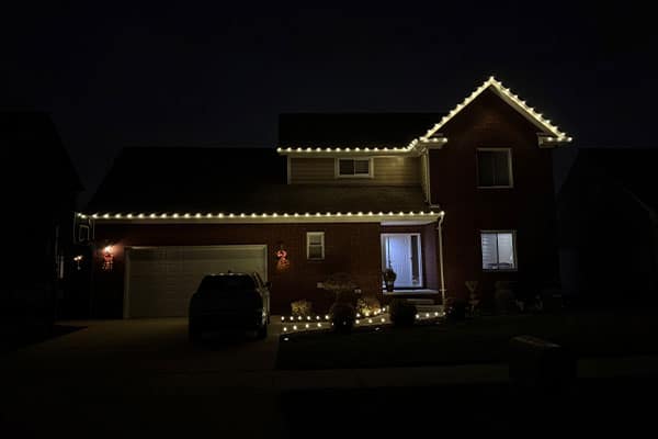 A suburban house at night, warmly lit with white Christmas lights outlining the roof and landscaping. A dark SUV sits in the driveway. Festive holiday atmosphere.