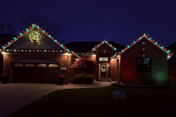 House decorated with red and green Christmas lights along the roofline, wreath on the garage. Sign in yard says "Christmas Light Installation.