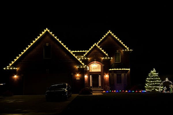 A brick house is adorned with warm white Christmas lights along the roofline. A decorated Christmas tree glows in the yard, with colorful lights lining the lawn. A car sits in the driveway.