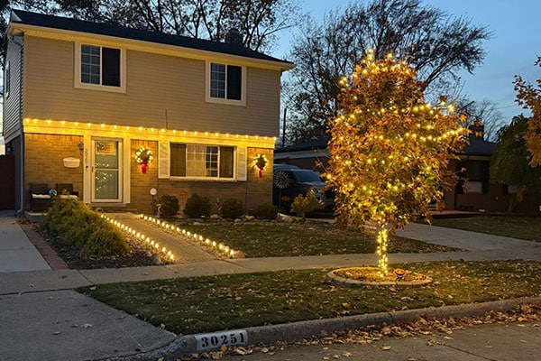 Two-story house glows with warm lights and festive wreaths. Path lined with lights leads to the door, and a tree covered in lights stands in the front yard, adding to the welcoming holiday ambiance.