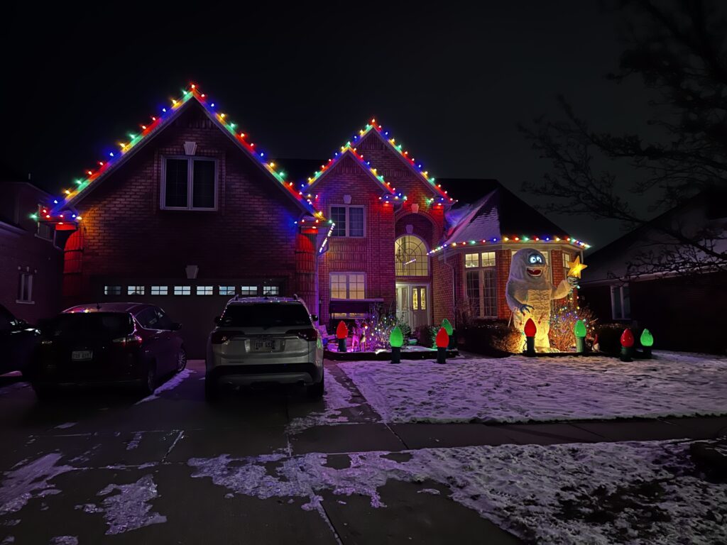 Festive house with Christmas lights outlining the roof. A large snowman decoration stands on the snowy lawn, near colorful light bulbs, after snow removal. Cars are parked in the driveway.